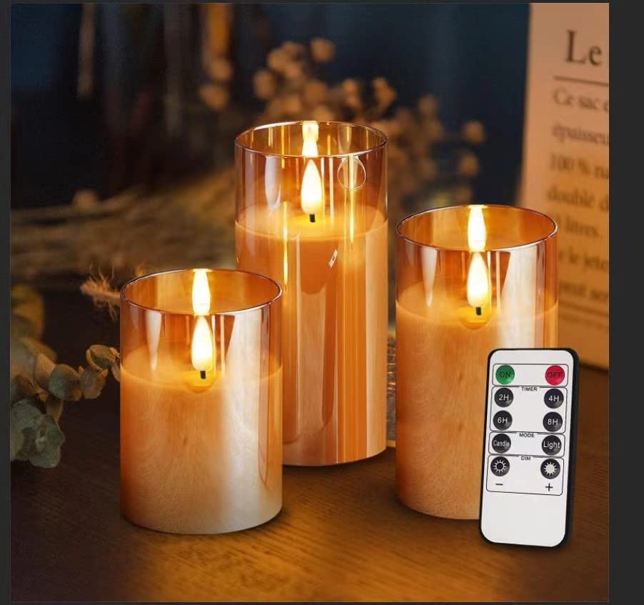 3 round glass containers of 3 different heights and of orange color with a candle in each one. The candles have flames. A flat, small remote control in a corner shows that these are non real candles but rechargeable ones