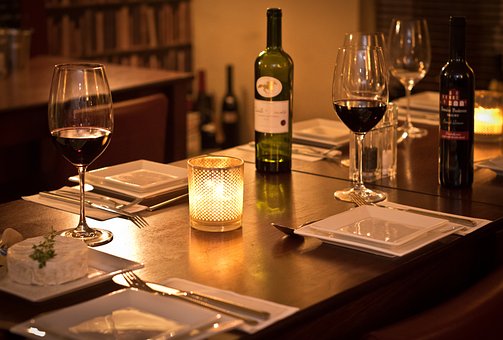 an illuminated candle in the center of a table dressed for a meal with square white plates and tall wine glasses filled with red wine. A bottle of wine on the side. A soft glow all over the photo.