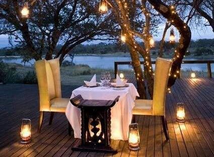 Table dressed for a meal at night, with 2 high back chairs, lanterns with illuminated candles on the floor round the table, candles on a trees nearby, garlands of lights in the tree and a candle on the table
