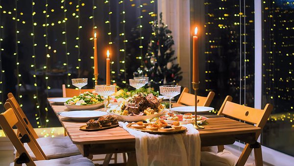 Dinner table set for 4 with 3 tapers candles lit on the table. The candles are orange. There are trays for food on the table and 5 champagne glasses. Garlands of lights hang on the wall behind the table and reflect in the window panels on the other side of the table.