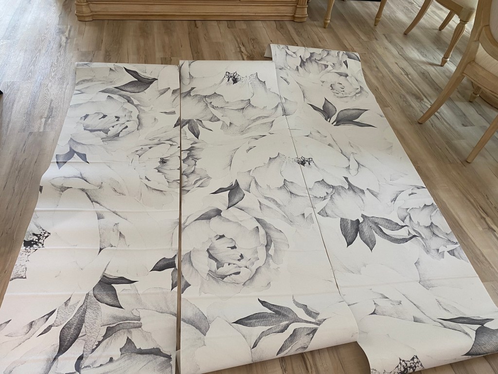 Three rolls of wallpaper printed with black and white p watercolors of peonies. The three rolls are displayed side by side on a wooden floor.
